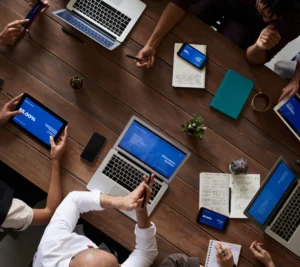 Group of people at a wooden table reviewing presentations on laptops, tablets, and handwritten notes