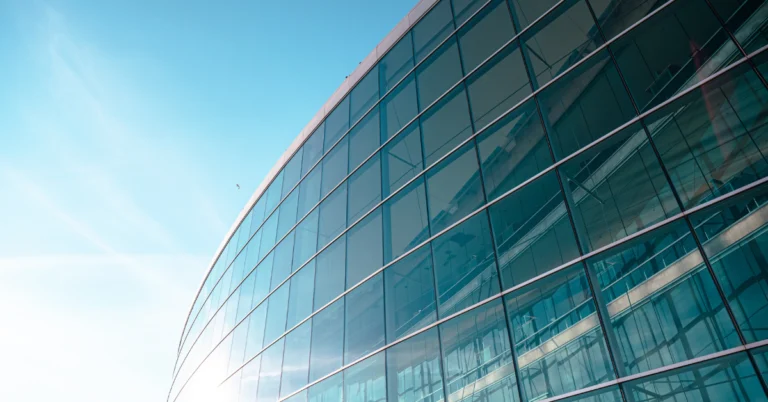 Modern glass office building reflecting the sky and nearby structures, photographed from a low angle against a bright blue sky, conveying a corporate or financial institution setting.