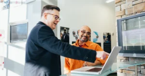 Two men collaborating at an office counter, one wearing a dark blazer and pointing at a laptop screen while the other, in an orange shirt, smiles and listens attentively.
