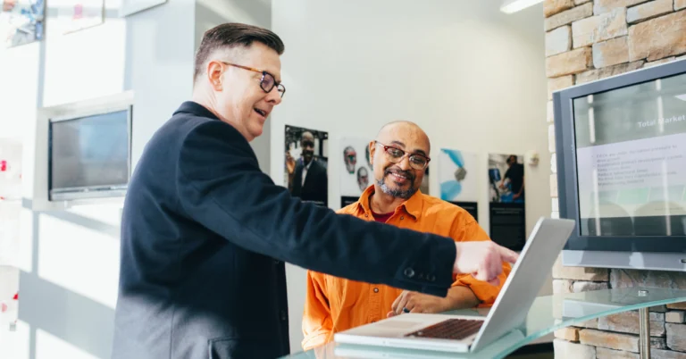 Two men collaborating at an office counter, one wearing a dark blazer and pointing at a laptop screen while the other, in an orange shirt, smiles and listens attentively.