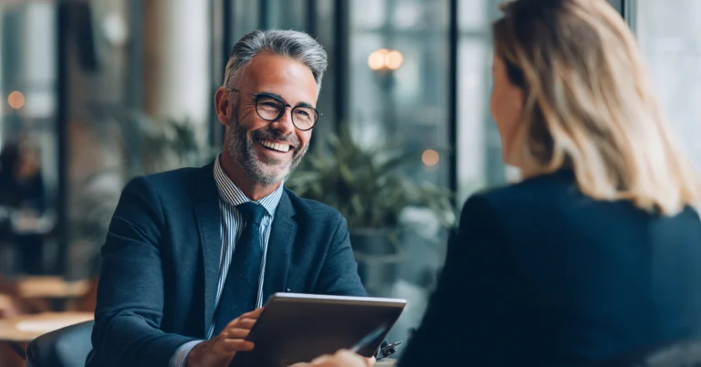 A cheerful man in a suit and glasses holds a tablet while talking with a colleague in a professional office setting.