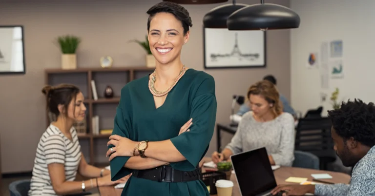 Smiling businesswoman standing confidently in a modern office with arms crossed, while her team works in the background with laptops and documents.