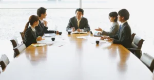 Business team in formal attire holding a meeting around a large conference table, discussing reports and project plans in a bright office setting.