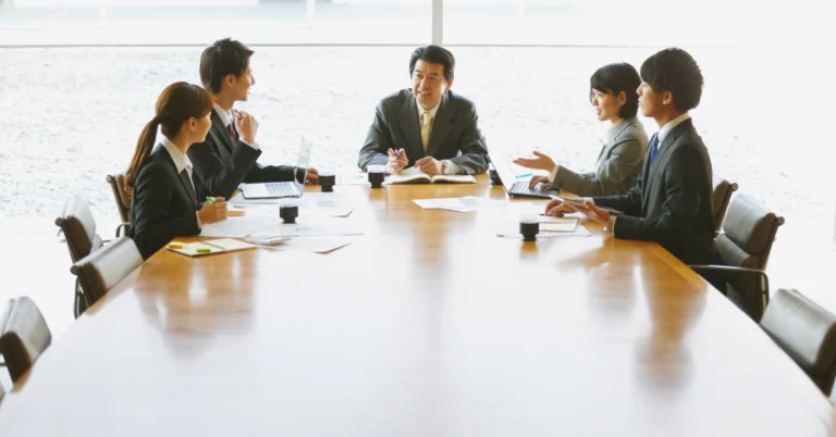 Business team in formal attire holding a meeting around a large conference table, discussing reports and project plans in a bright office setting.