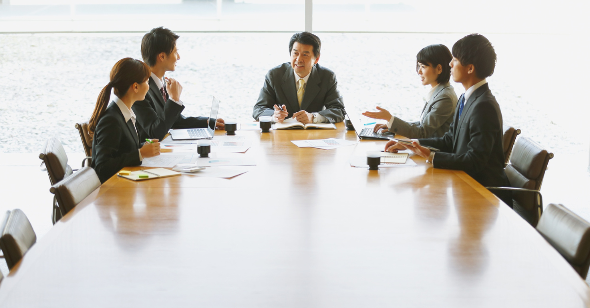 Business team in formal attire holding a meeting around a large conference table, discussing reports and project plans in a bright office setting.
