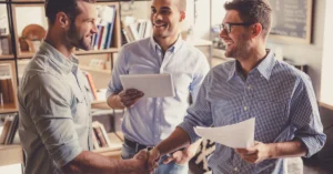 Smiling colleagues shaking hands while another person holds a tablet in the background, symbolizing teamwork and business agreement in a casual office environment.