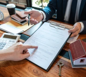 A person hands over a pen while another points at a document on a clipboard, with a calculator, notebook, and small house model on the desk.