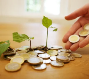 Several small plants sprout from piles of coins on a wooden table while a hand places additional coins beside them.