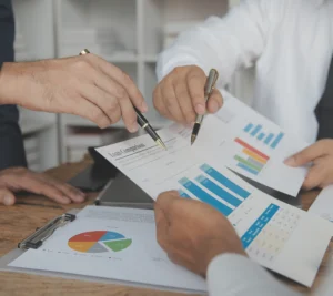 Close-up of three people reviewing financial charts and reports with pens, discussing bar graphs and pie charts on printed documents during a business meeting.
