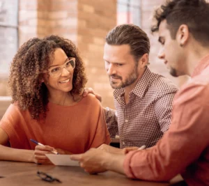 A smiling woman with curly hair and glasses talks with two men at a table, all reviewing a document together in a warm, casual setting.