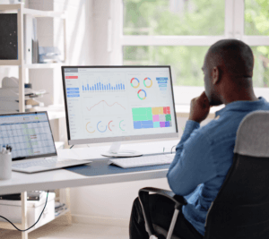 A man sitting at a desk analyzing dashboards and charts displayed on a large computer monitor and a laptop.